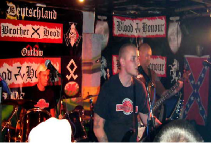 Konzertszene von 2006. Nach dem Verbot von Blood & Honour in Deutschland bildeten sich Nachfolgestrukturen, die sich u.a. Division 28 und Brotherhood 28 nannten. Auf dem Foto ist ein Bühnentransparent der Brotherhood 28 zu sehen.  In der Bühnendekoration sind zudem die Odal-Rune und die Südstaatenfahne zu erkennen.