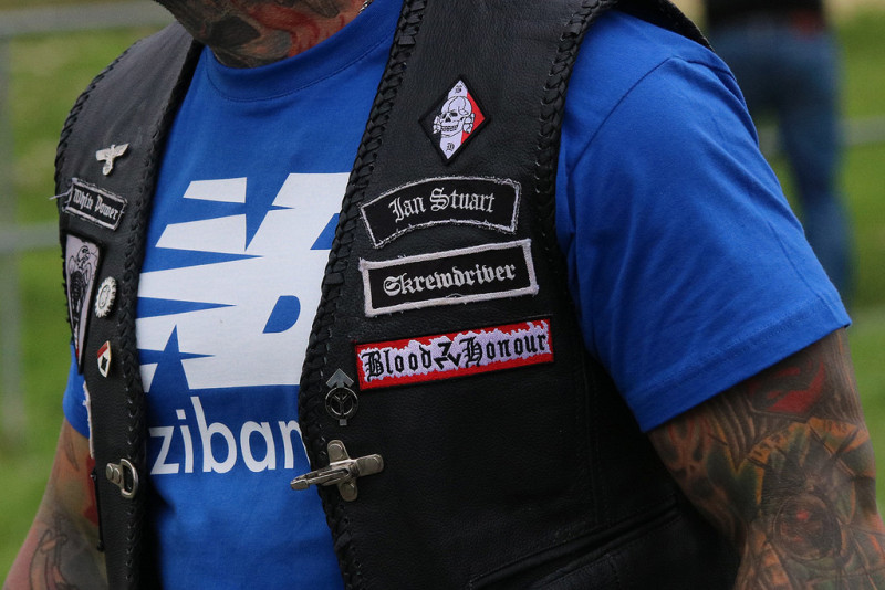 Aufnäher mit SS-Totenkopf und dem Organisationskennzeichen von Blood & Honour, gezeigt auf einem Neonazikonzert 2017 in Thüringen. Beide Symbole sind in dieser Darstellung verboten. Auf der Weste sind weiterhin zu erkennen: Die Tyr-Rune und die Lebensrune, das Zahnrad, der Reichsadler sowie ein White-Power-Slogan. Das Shirt-Motiv zeigt das NB-Logo der populären Bekleidungsmarke New Balance - allerdings als rechte Nachahmung, in der NB für »Naziband« steht.
