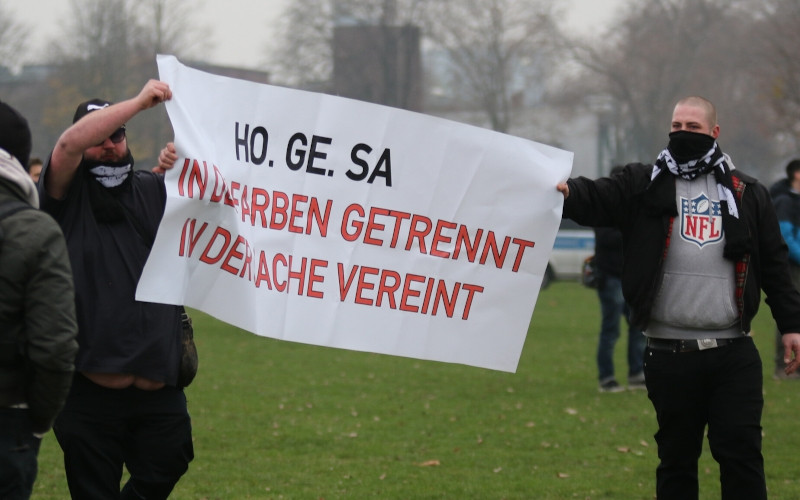 Transparent auf der Versammlung von Corona-Leugner:innen am 6. Dezember 2020 in Düsseldorf. Das Zeigen dieses Transparents weist nicht auf eine strukturelle Wiederbelebung von HoGeSa hin sondern es ist eher eine Erinnerung an vergangene HoGeSa-Zeiten. Die Parole »In den Farben getrennt in der Sache vereint« drückt aus, dass bei HoGeSa Fans unterschiedlicher Fußballvereine (Farben), die ansonsten verfeindet sind, politisch gemeinsam agieren.